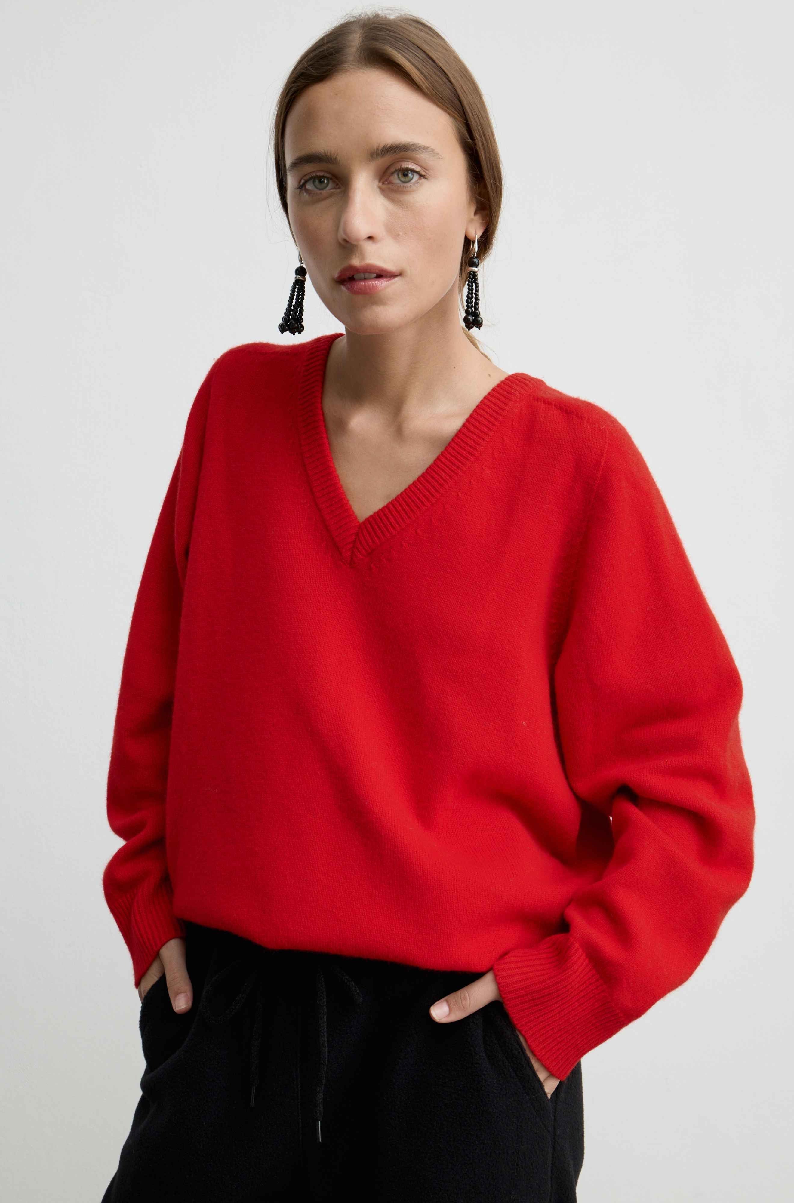 A woman wearing a bright red V-neck knit sweater and black pants, standing against a light gray studio background.