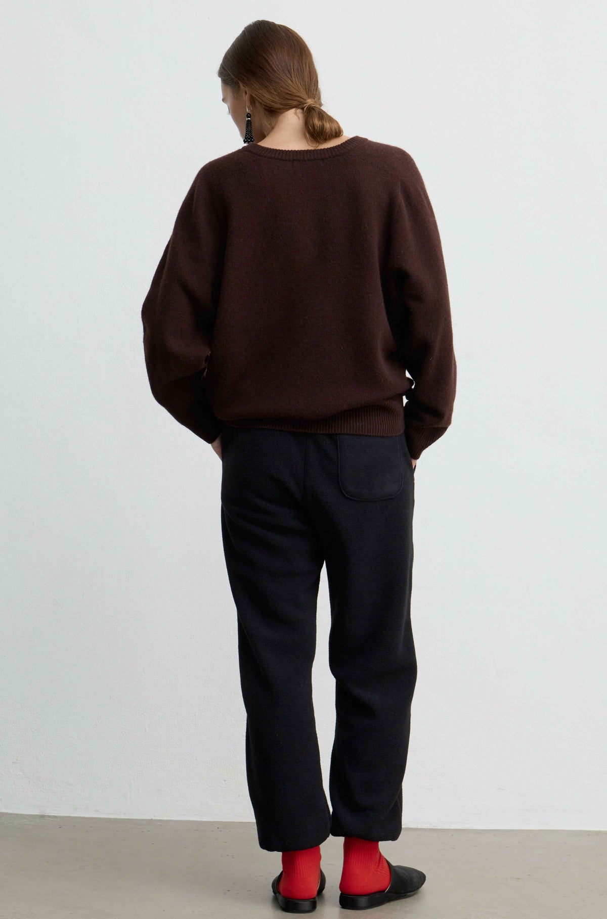 A person shown from the back wearing a dark brown knit sweater, black trousers, and bright red socks against a plain light gray studio background.
