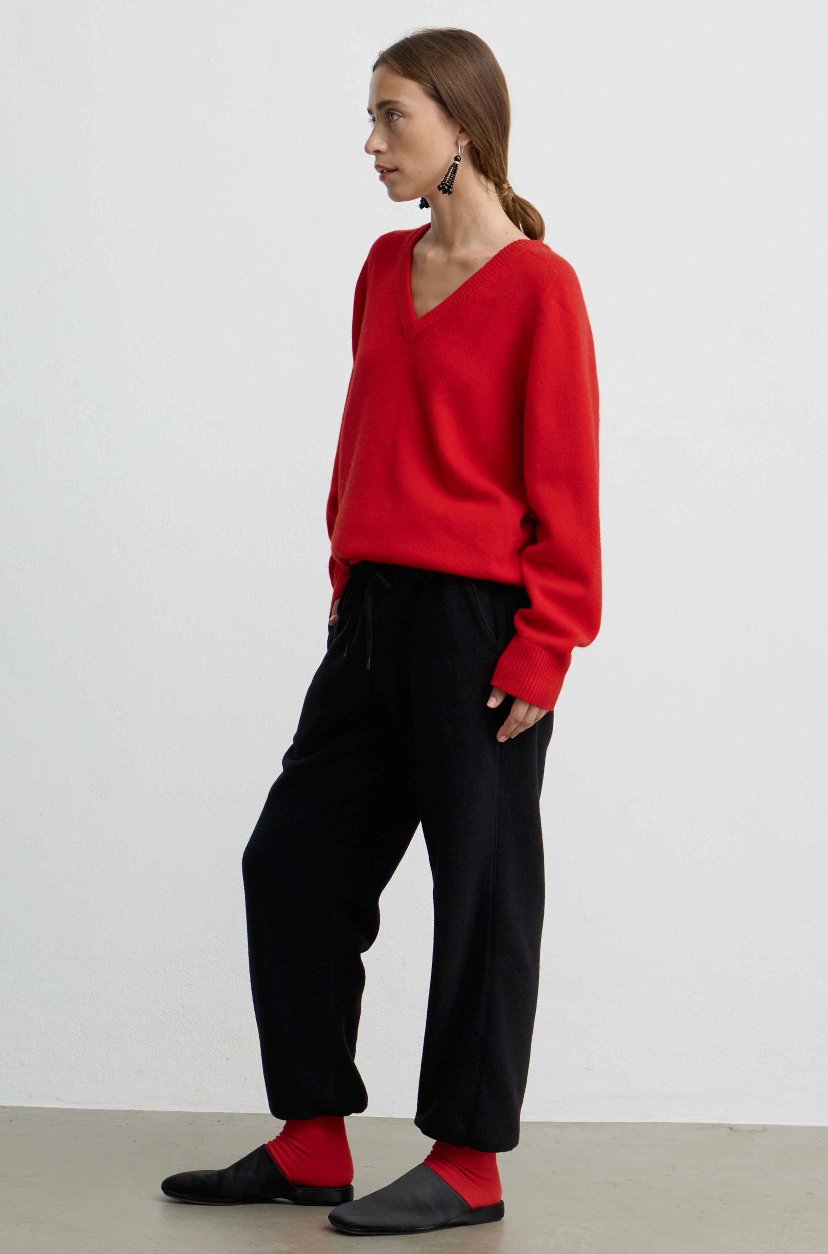 A woman stands in profile wearing a bright red V-neck knit sweater, black wide-leg pants, red socks, and black slip-on shoes against a plain light gray studio background.