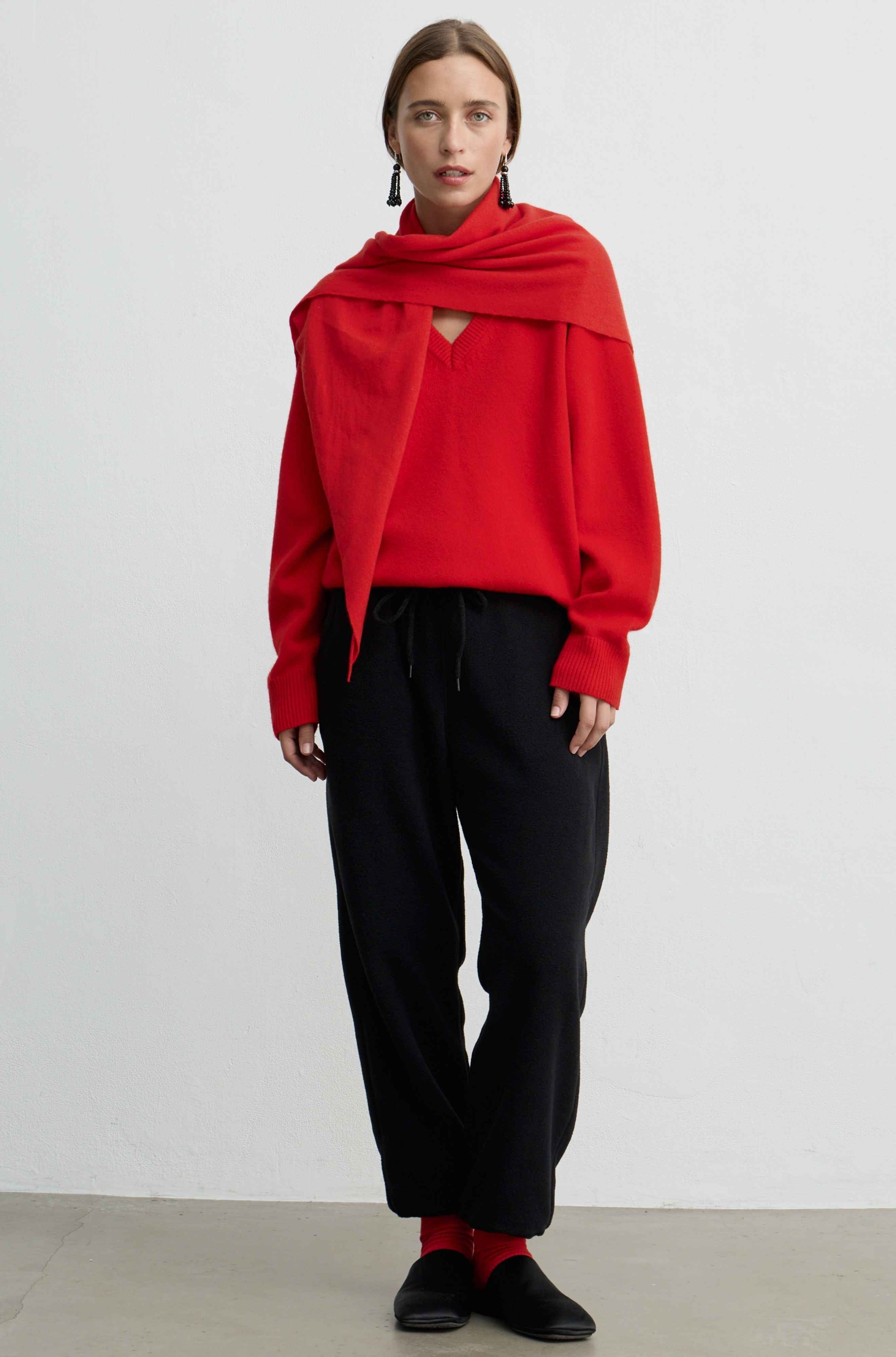 A model stands against a plain light gray wall wearing a bright red draped knit top with a keyhole cutout, black drawstring trousers, black loafers, and red socks.