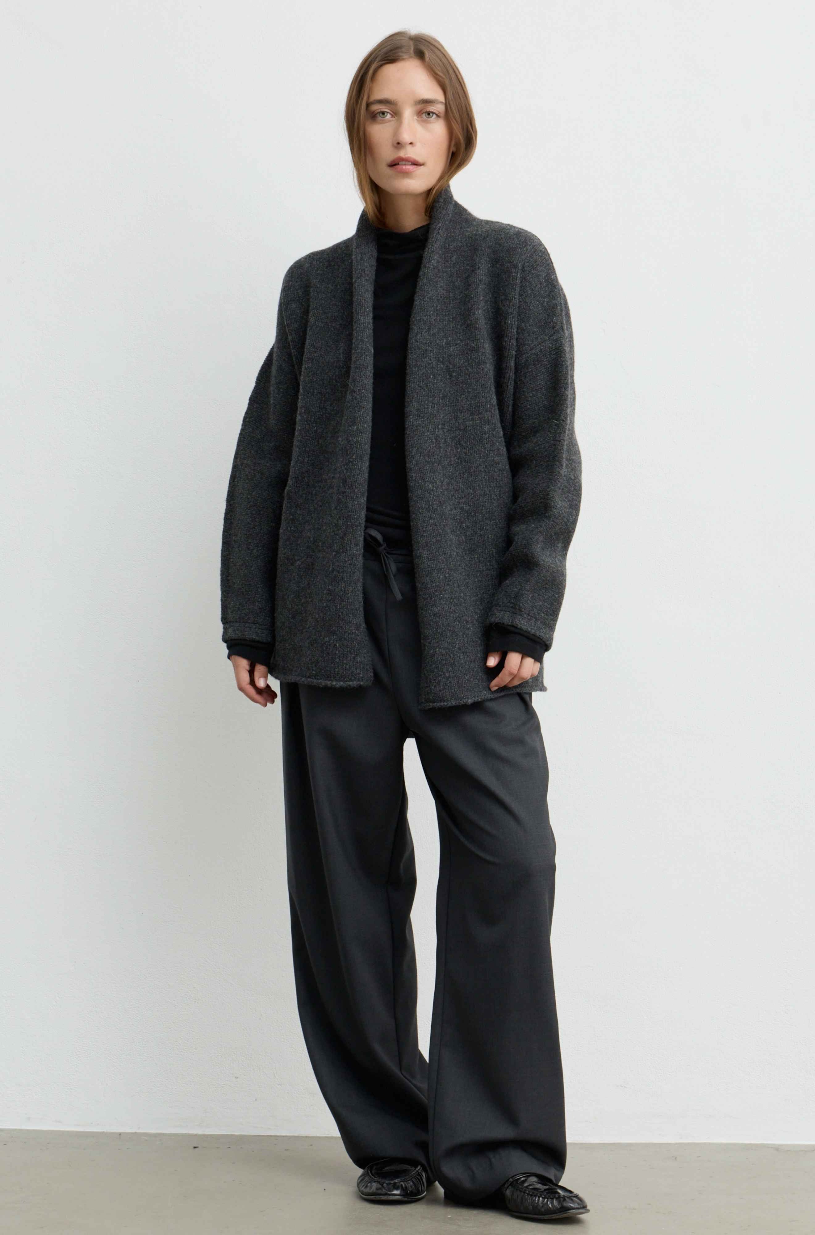 A model stands against a plain light gray wall wearing a dark charcoal gray open-front cardigan over a black turtleneck, paired with wide-leg dark trousers and black shoes.
