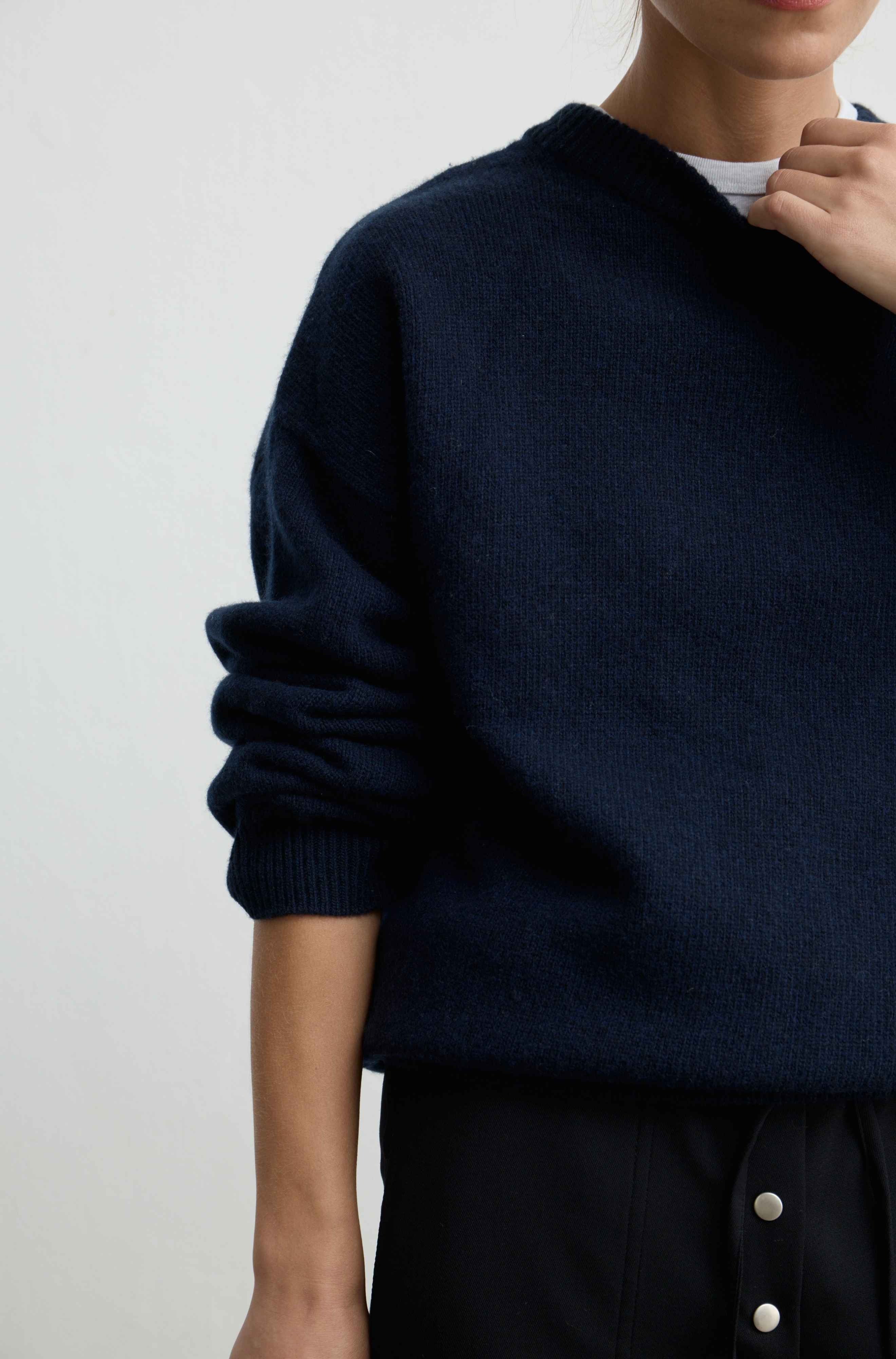 A close-up of a person wearing a dark navy knit sweater with rolled sleeves, layered over a white undershirt and paired with black button-front bottoms, against a plain light background.