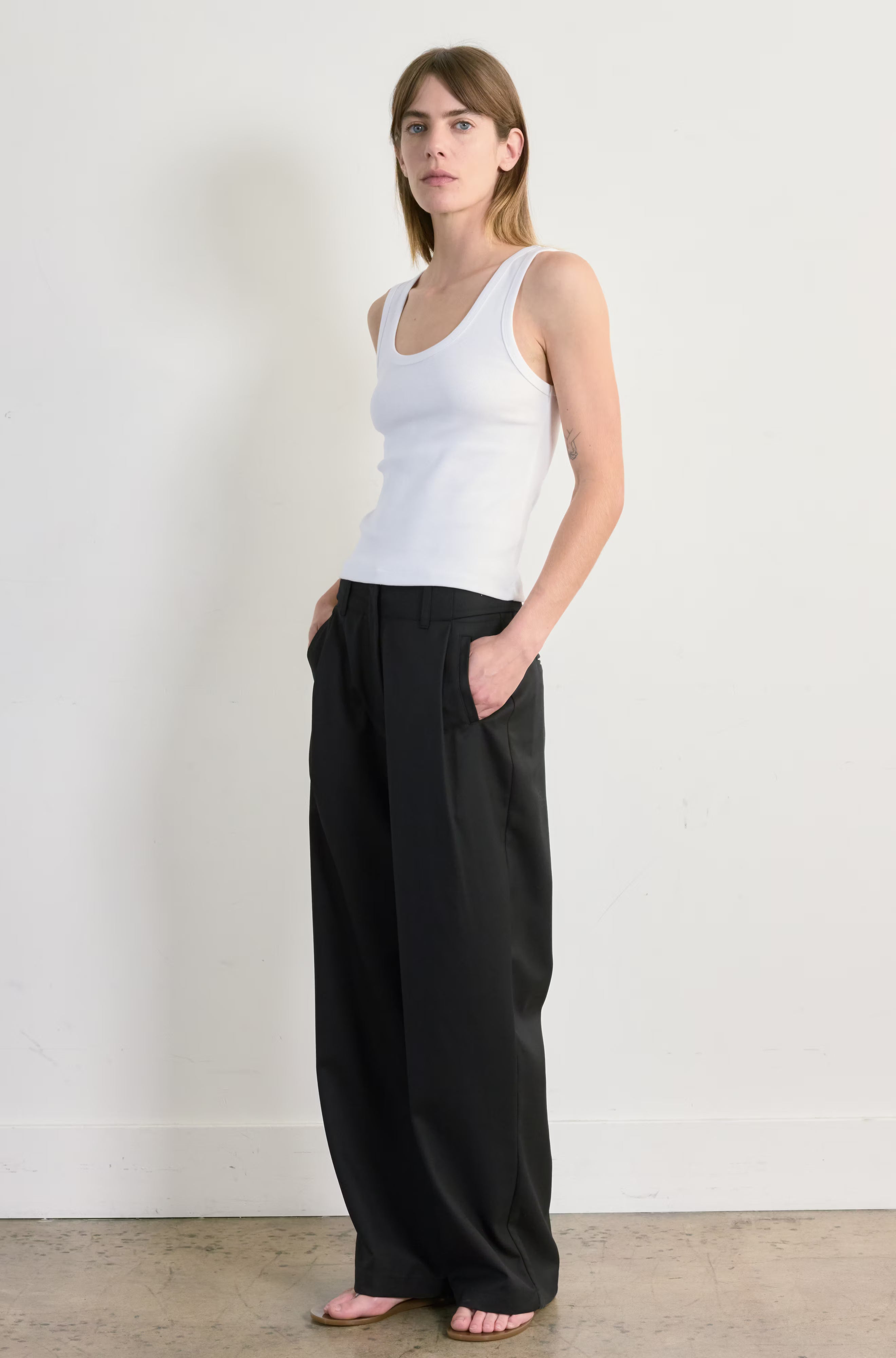 Woman wearing a white tank top and black pants against a plain background