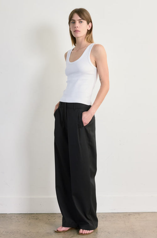 Woman wearing a white tank top and black pants against a plain background