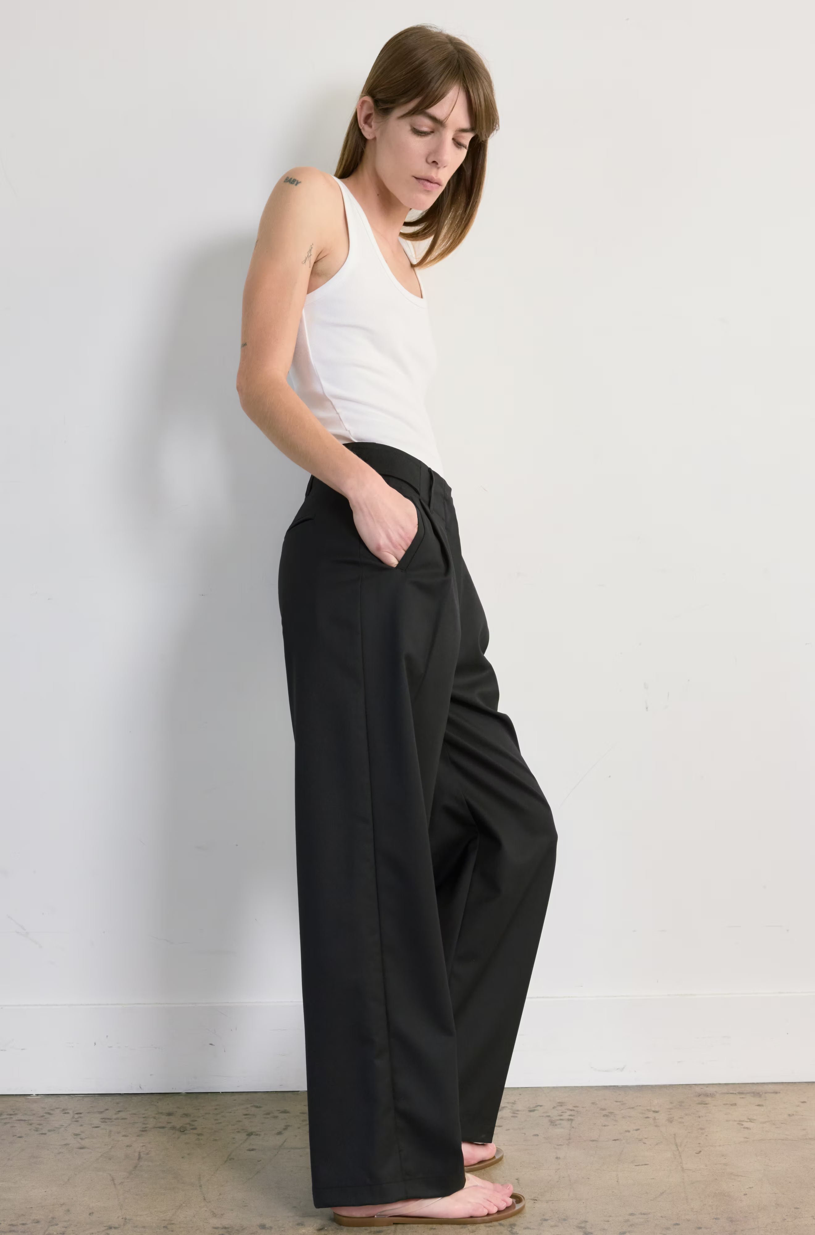 Woman wearing a white tank top and black wide-leg pants against a plain white wall.
