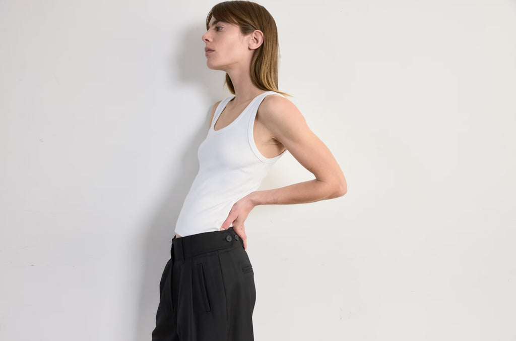 Woman wearing a white tank top and black pants against a plain background