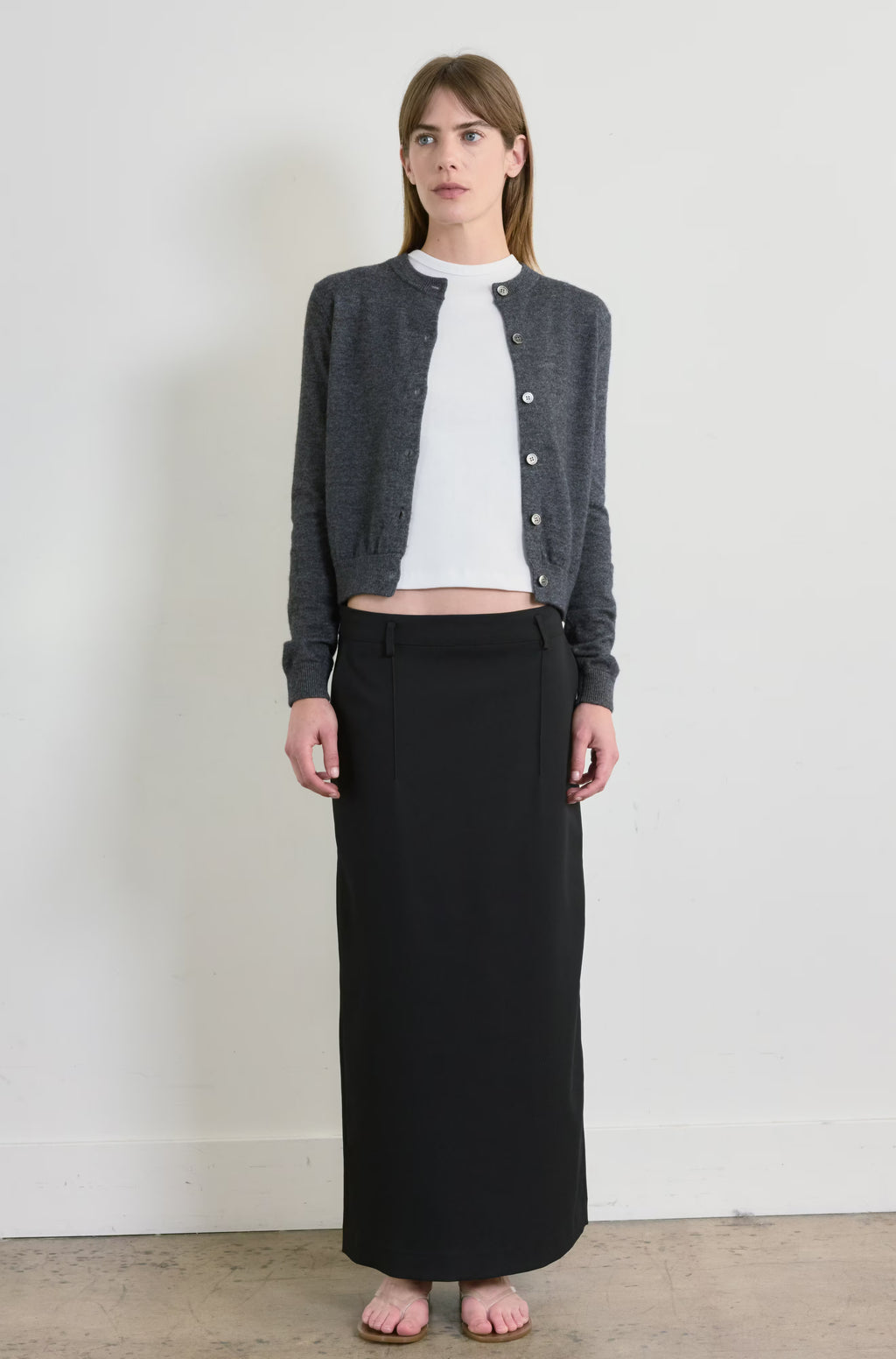Woman wearing a gray cardigan, white shirt, and black skirt against a plain background