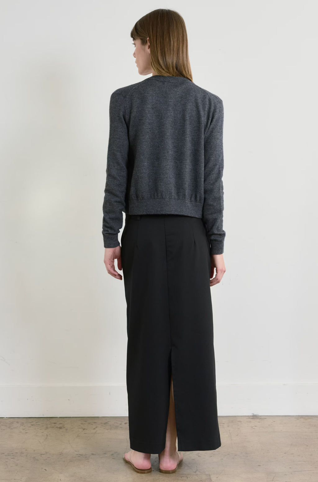 Person wearing a dark gray cardigan and black skirt against a white wall.