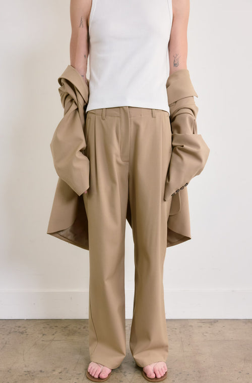 Person wearing beige pants and a white top, holding a beige jacket against a plain background