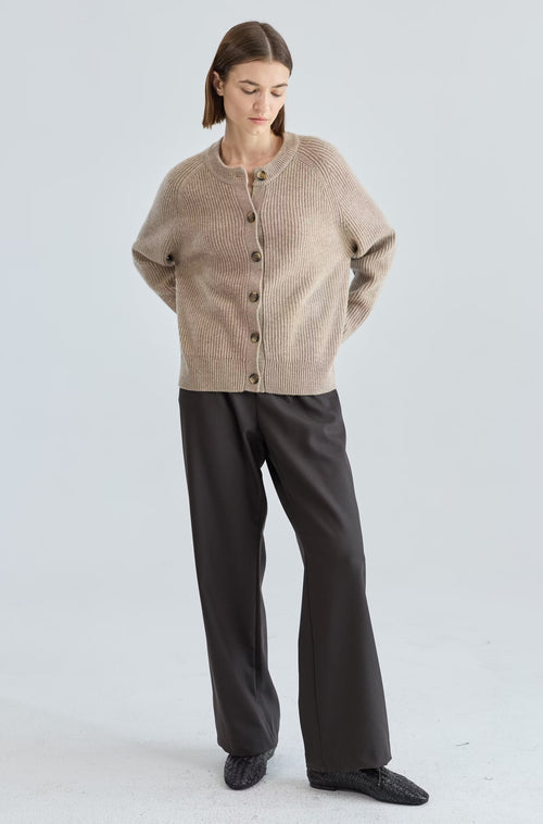 Woman wearing a beige cardigan and dark pants on a light gray background