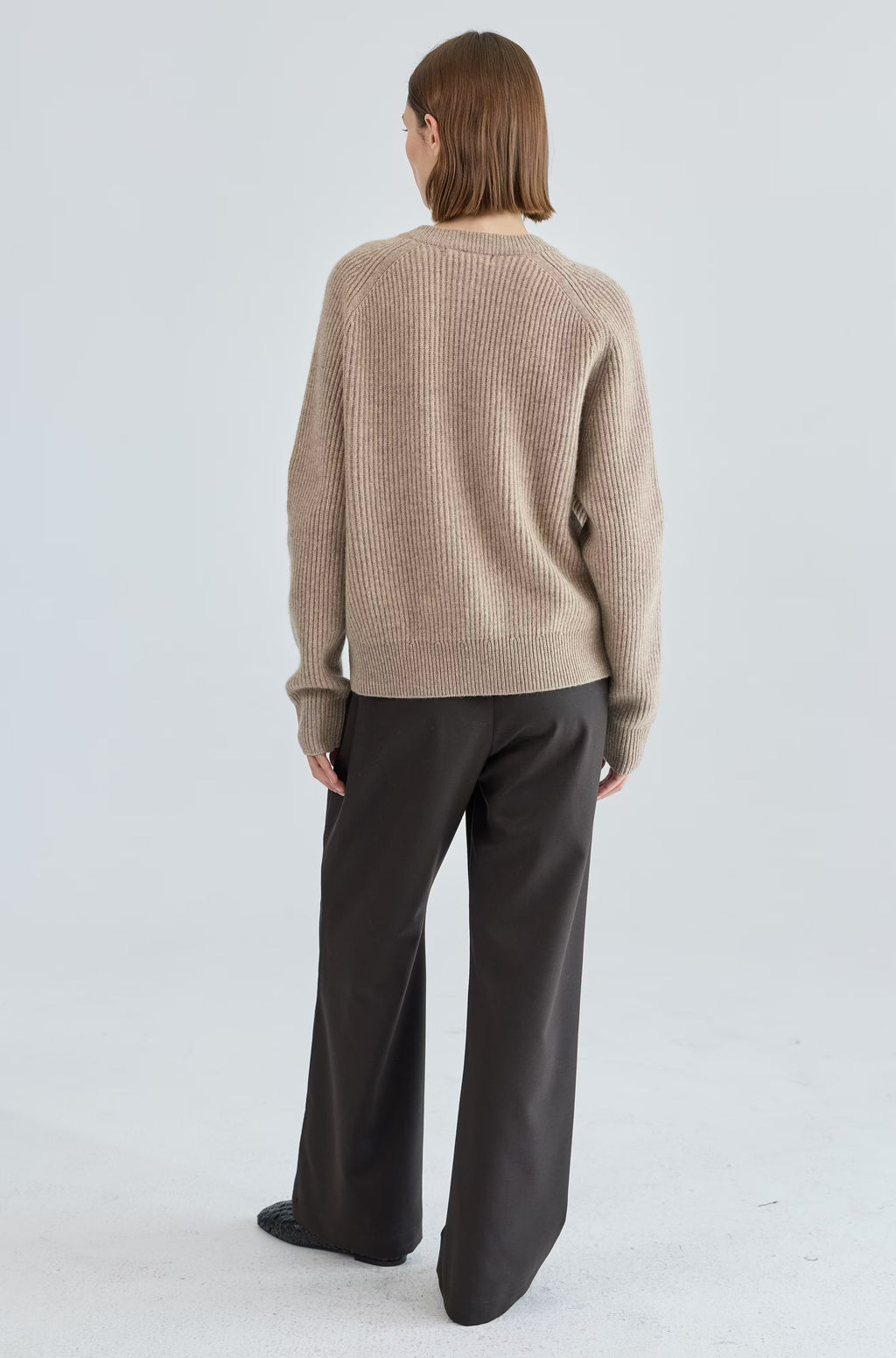 Person wearing a beige sweater and dark pants on a light gray background