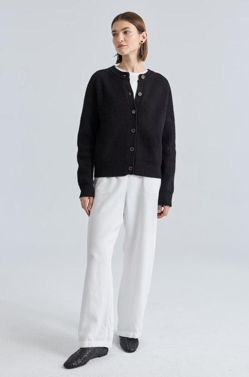 Woman wearing a black cardigan, white shirt, and white pants on a light gray background