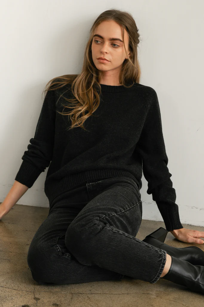 Woman wearing a black sweater and jeans sitting against a white wall.