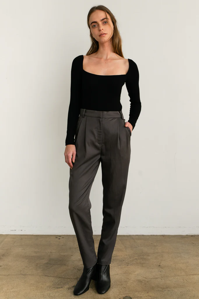Woman wearing a black top and gray pants against a white wall.