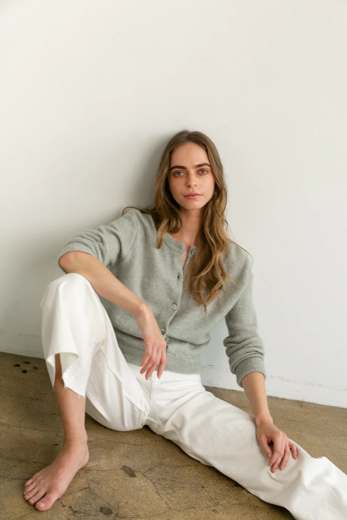 Woman sitting on the floor wearing a gray button up cardigan and white pants against a plain wall.
