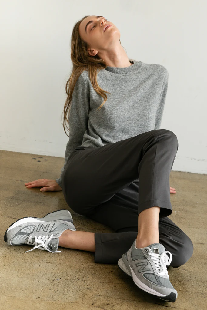 Woman sitting on the floor wearing a gray sweater, black pants, and gray sneakers.