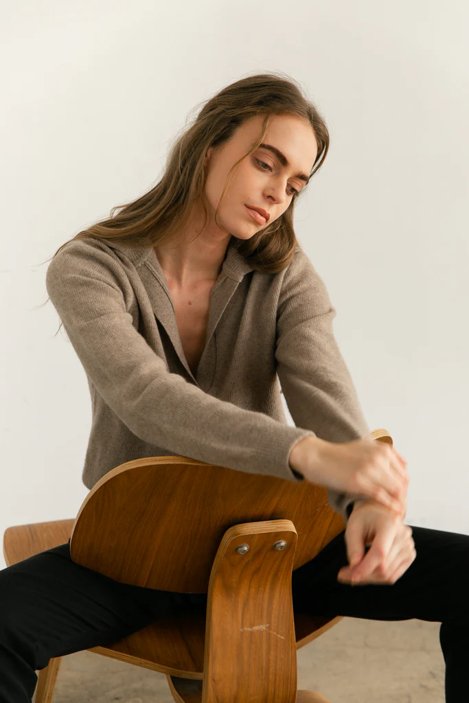 Woman sitting on a wooden chair against a plain background. Wearing a V Neck Wool Sweater in beige.
