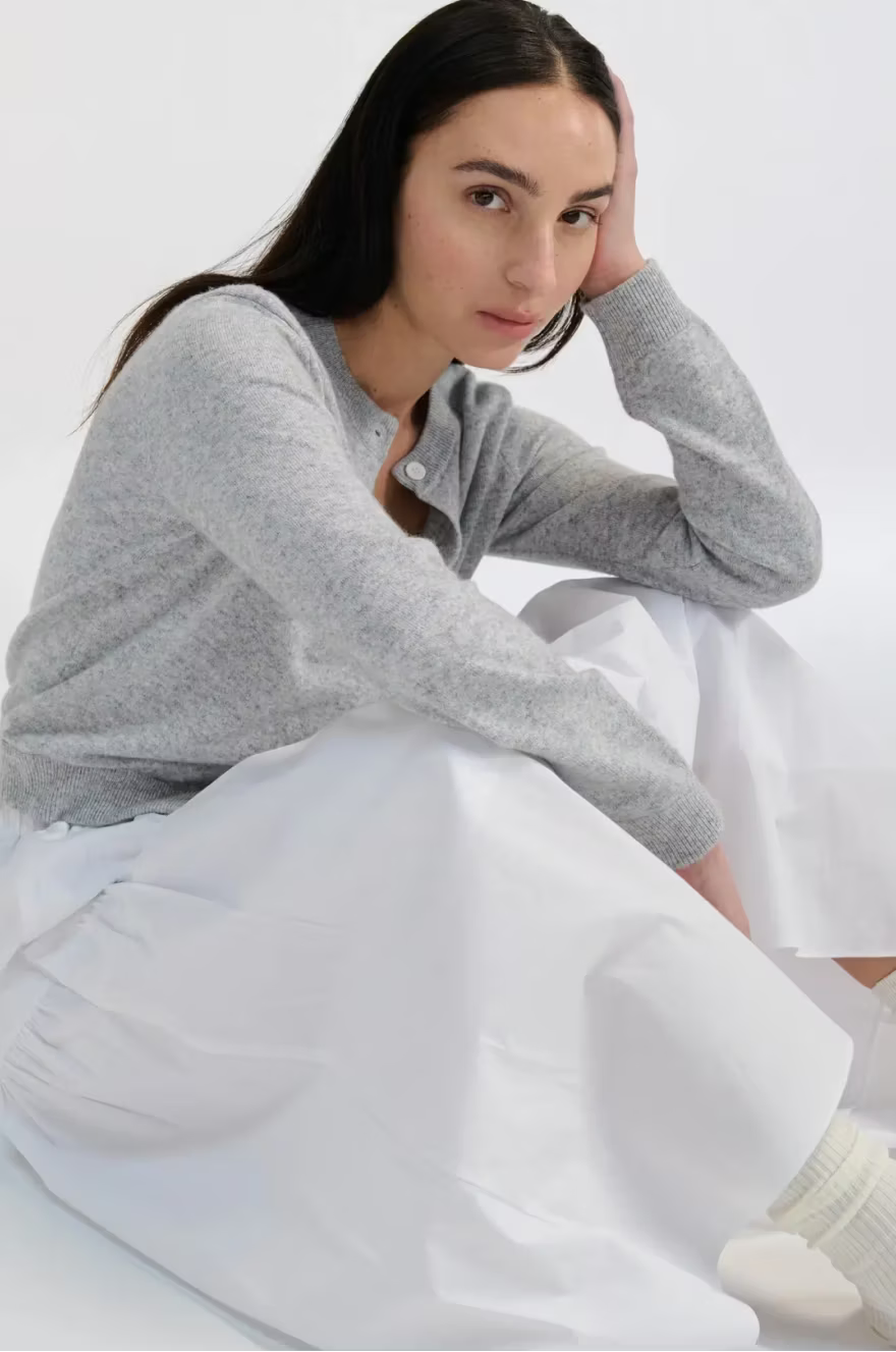 Woman wearing a gray sweater and a white skirt sitting on a white surface.
